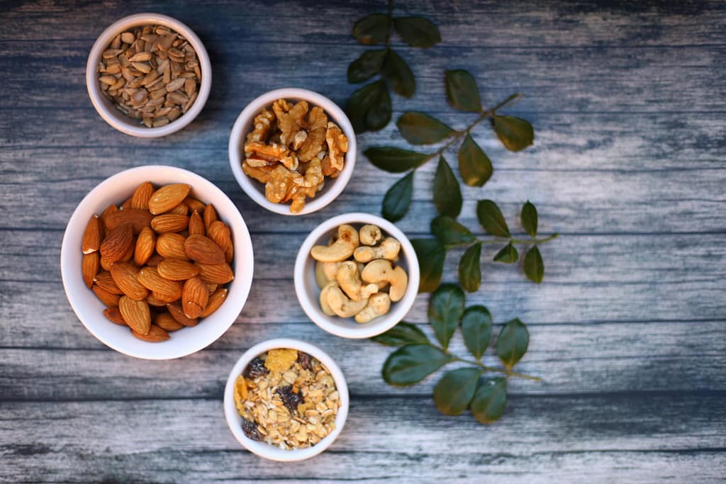 Aerial view of assorted nuts and seeds in white bowls with green leaves on a rustic wooden table.